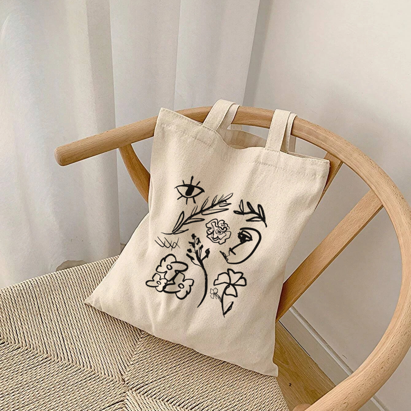 Beige tote bag with black floral and eye designs on a wooden chair against a white curtain background